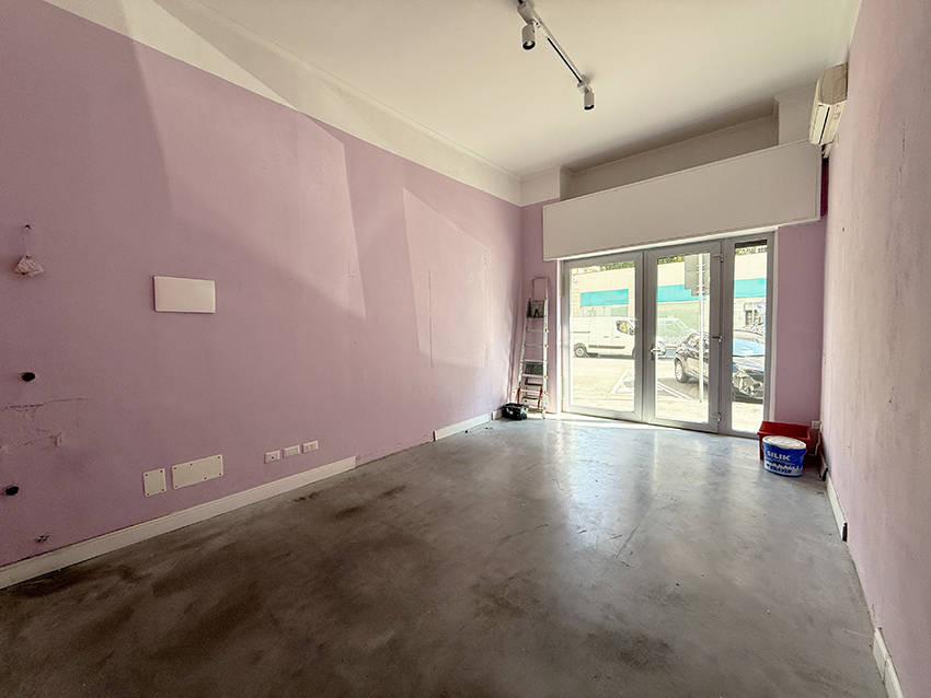 Interno del negozio in affitto in via Negroli a Milano: locale vuoto con pareti lilla, pavimento grigio, binario con faretti sul soffitto, climatizzatore e vetrina sulla strada sul fondo.
