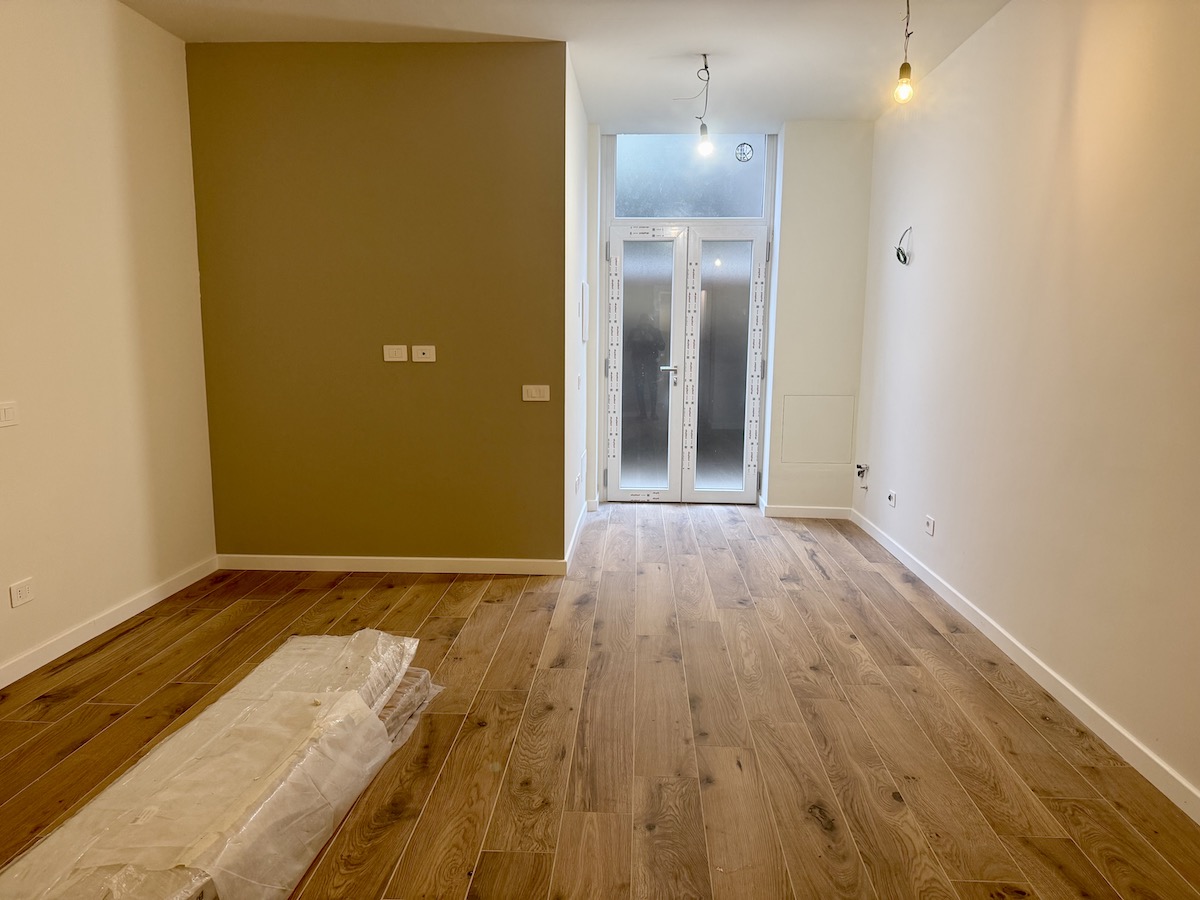 Monolocale ristrutturato in vendita a Milano in via Tajani 10, zona Città Studi: ambiente vuoto con pavimento in legno chiaro, parete d'accento color tortora, pareti bianche, lampadine a sospensione e porta d'ingresso a due ante in vetro satinato sul fondo.