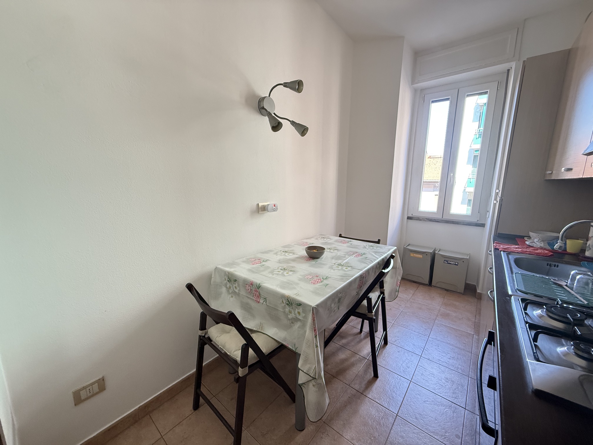 Cucina abitabile del bilocale in vendita a Milano in via Azzo Carbonera, zona Porta Vittoria: tavolo con tovaglia a fiori e due sedie pieghevoli in legno scuro, applique a due luci sulla parete bianca, pavimento in piastrelle color cotto e finestra in PVC con doppio vetro sul fondo.