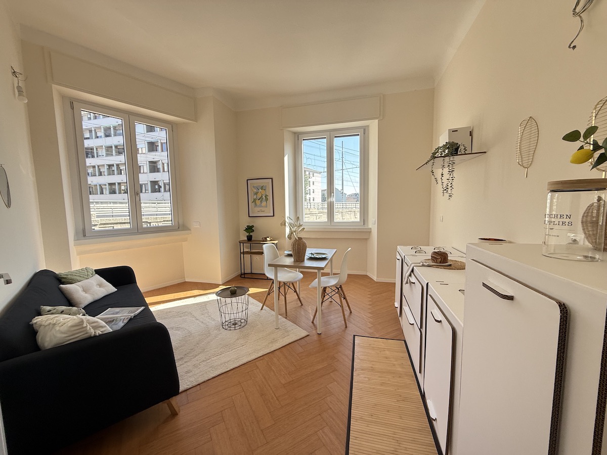 Zona giorno del bilocale ristrutturato in vendita a Milano in Viale Monza 119: divano blu scuro con cuscini chiari, tappeto bianco, tavolo con due sedie bianche, cucina a vista bianca sulla destra, parquet a spina di pesce e due finestre in PVC che illuminano la stanza.