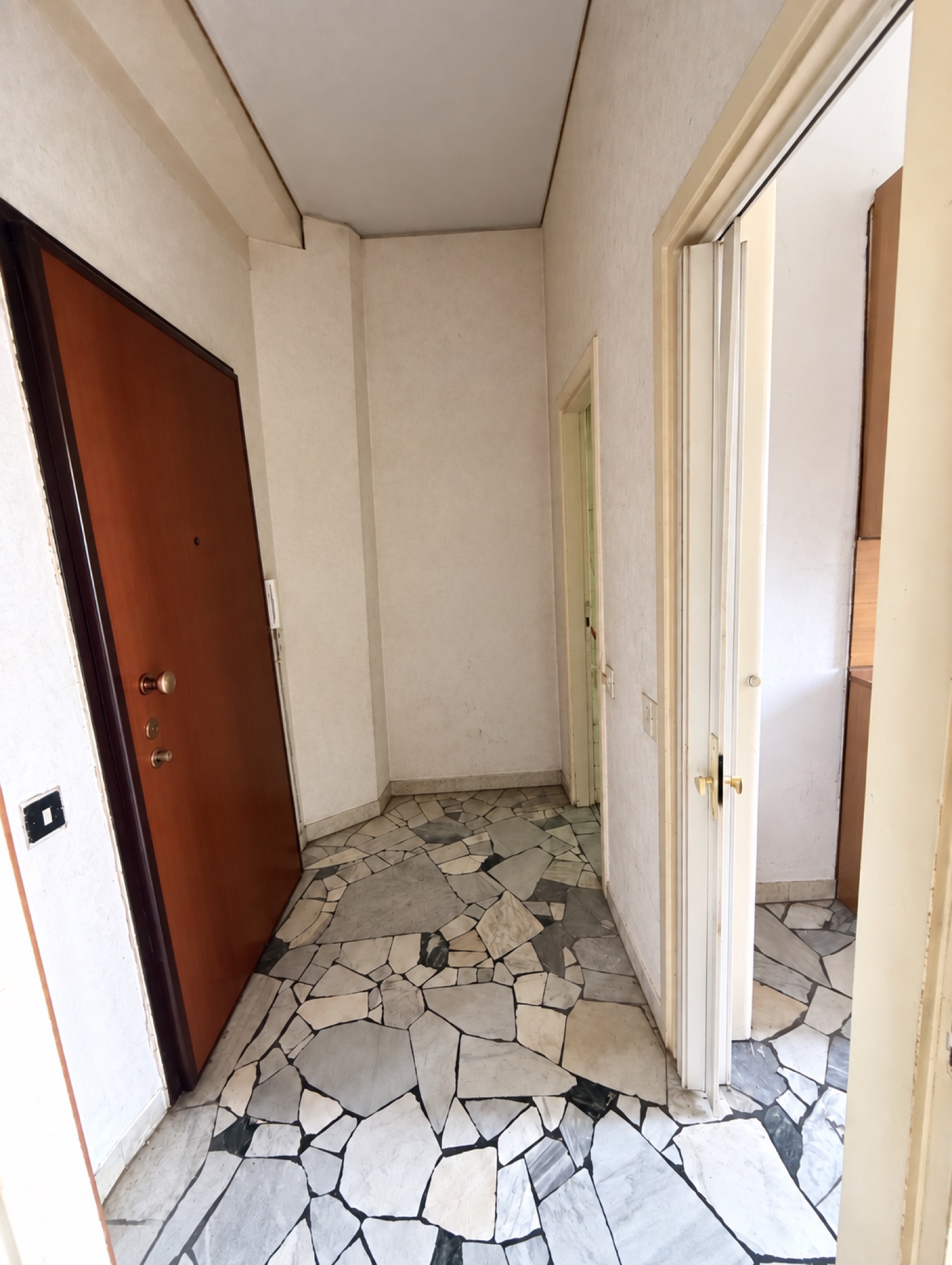 Ingresso del monolocale da ristrutturare in vendita a Milano in via Rogoredo 113: porta blindata in legno a sinistra, corridoio con pavimento in marmo a palladiana grigio e bianco, pareti bianche e porta aperta su una stanza a destra.